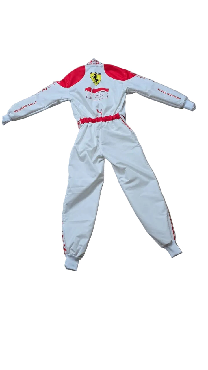 A white and red Scuderia Ferrari children's racing suit laid flat on a beige tiled floor. The suit features the Ferrari shield logo on the chest, red shoulder panels, a red waist belt, and red stripes on the sleeves and legs.